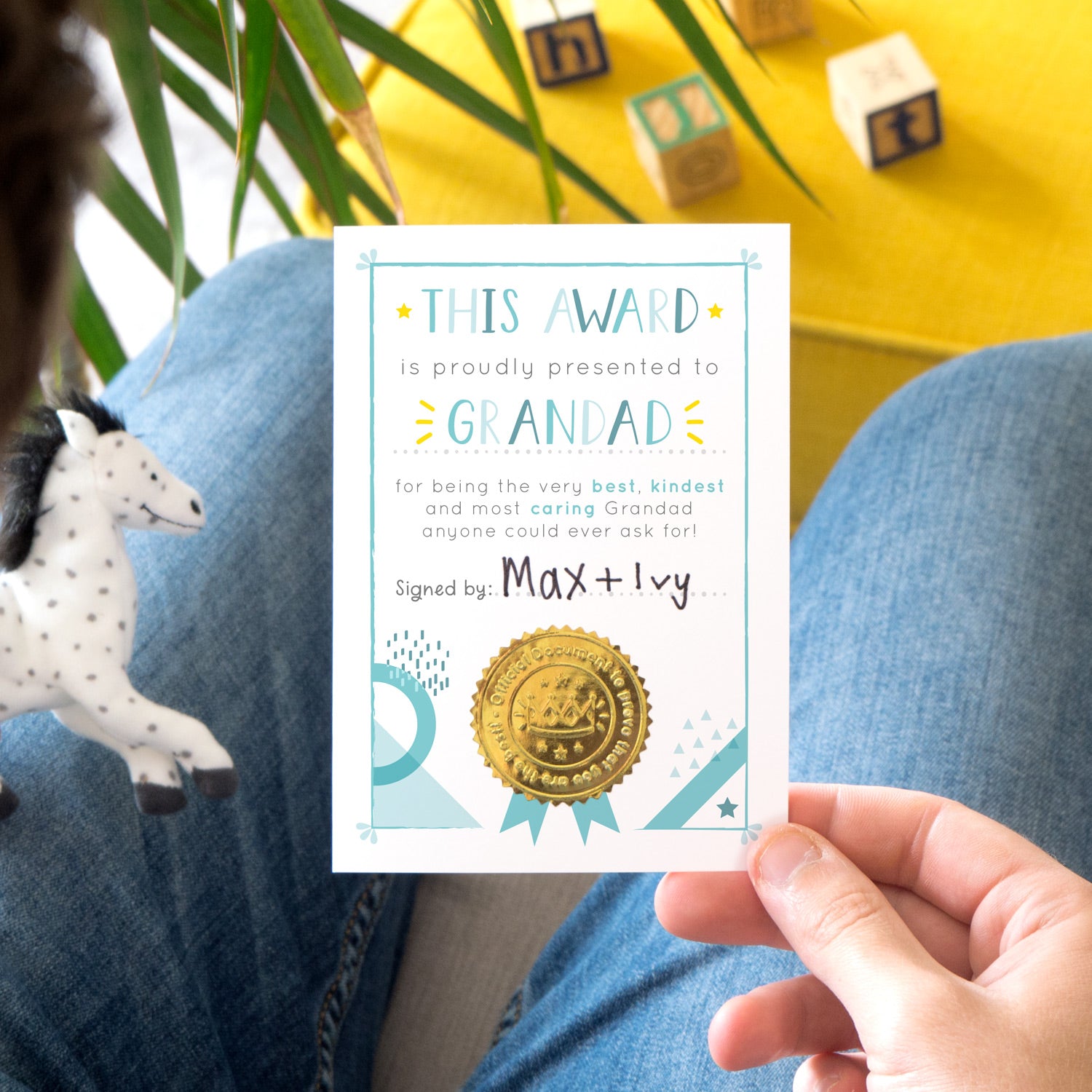 A Grandad certificate award card printed onto white card in varying tones of blue and pops of yellow! Each card features a shiny gold seal to make it official! Photographed over a lap with a yellow footrest in the background.