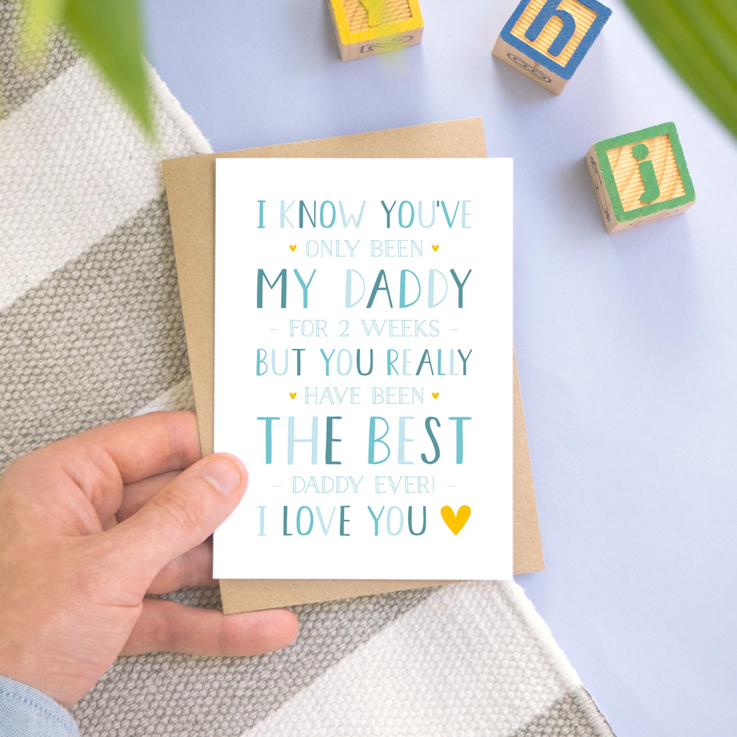 A personalised new daddy card photographed on a blue and grey stripy background with a few leaves, and wooden building blocks. The card is being held by a father and features typography in varying tones of blue and with a pop of yellow.