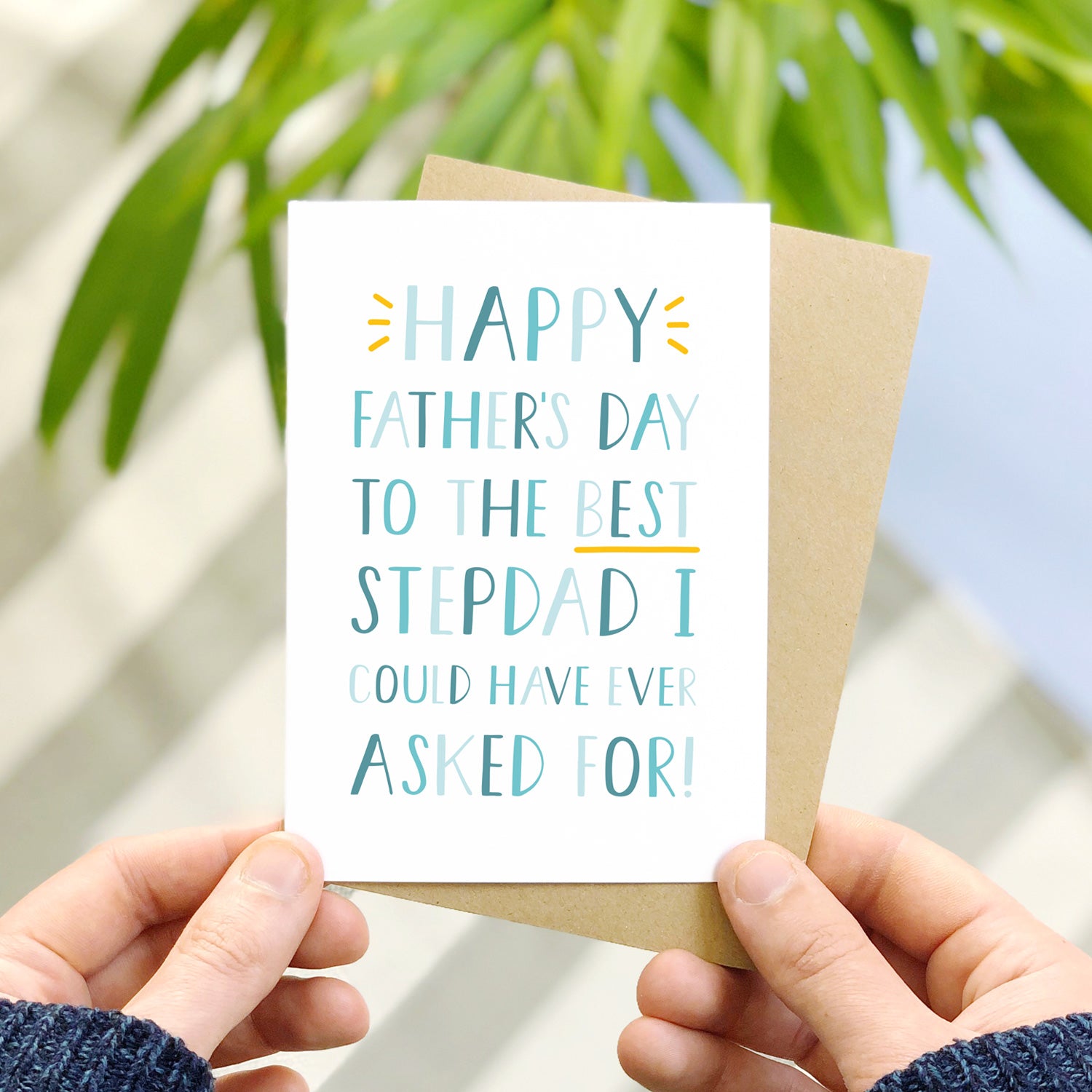 Happy Father's Day to the best stepdad I could have ever asked for Father's Day card. It has been photographed over a grey striped rug with a blue floor and some leafy foliage. It is also being held by a man.