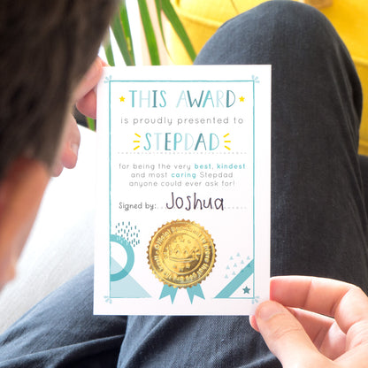 A stepdad award certificate card printed onto white card, with varying tones of blue and pops of yellow. This card is photographed being held over black jeans and a yellow footrest.