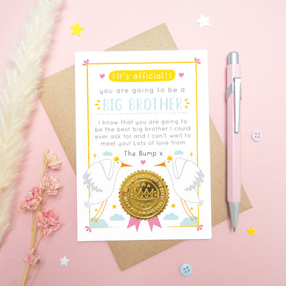 A big brother announcement card from the bump photographed on a pink background with dried flowers, a pen, buttons and stars.