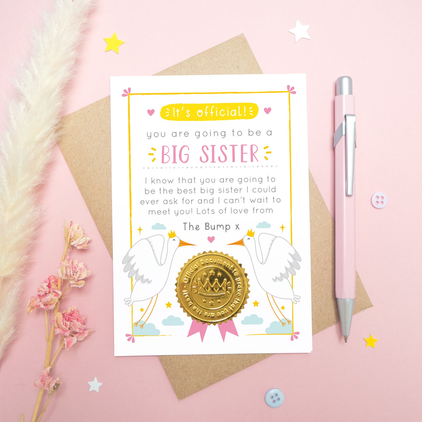 A big sister announcement card from the bump photographed on a pink background with dried flowers, a pen, buttons and stars.