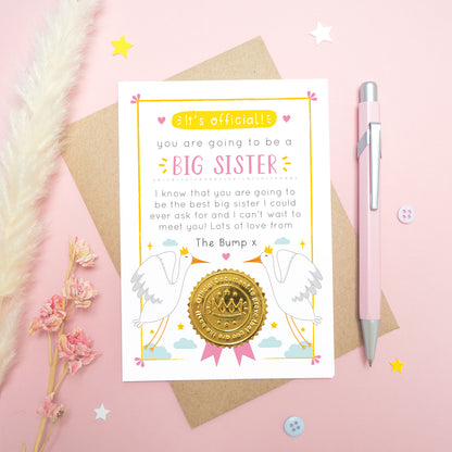 A big sister announcement card from the bump photographed on a pink background with dried flowers, a pen, buttons and stars.