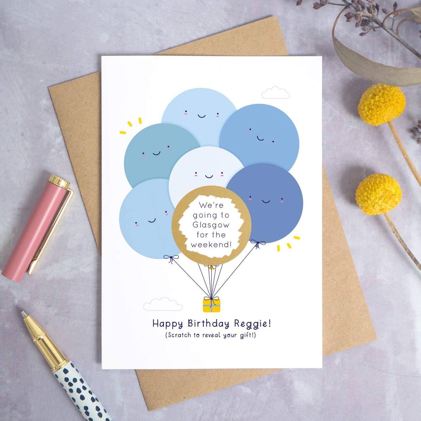 A ‘happy birthday’ scratch card photographed on a grey background with a pen on the left and yellow and purple flowers on the right. This is the blue version of the card after it has been scratched off.