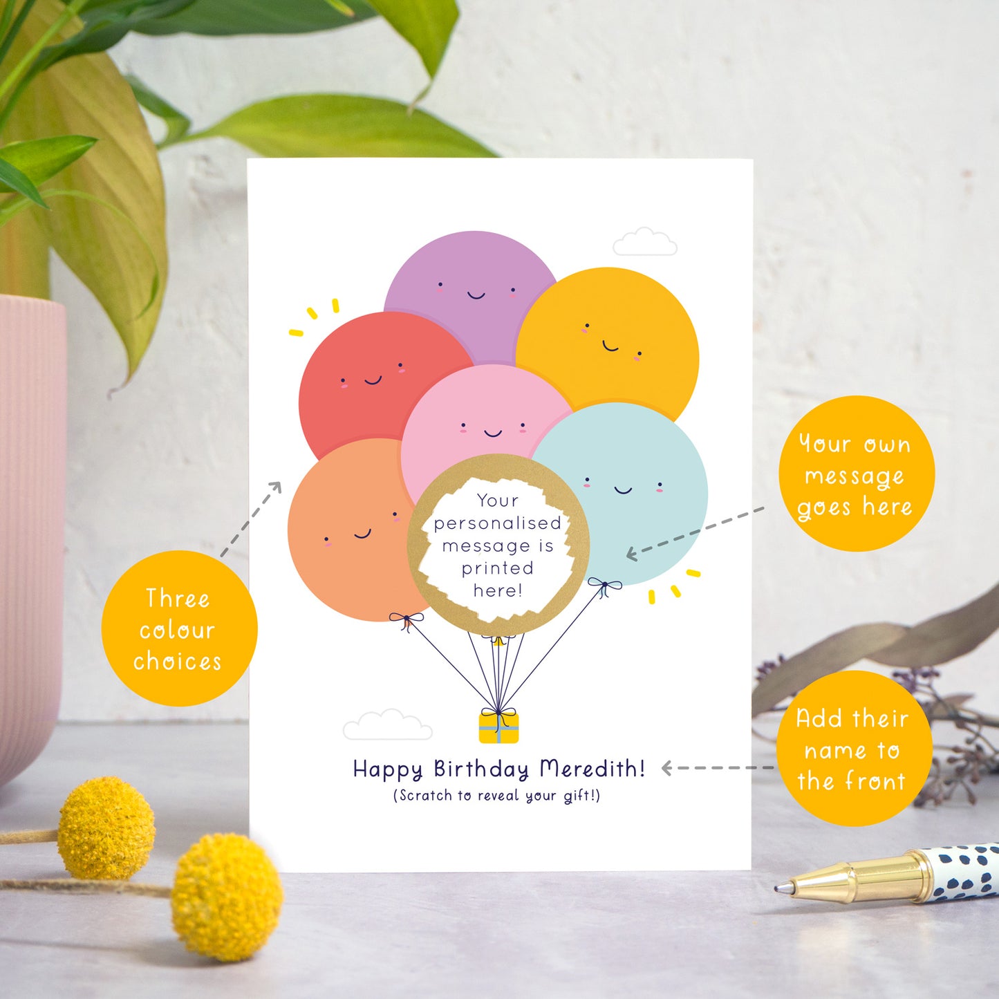 A ‘happy birthday’ scratch card photographed on a white and grey background with a plant on the left and foliage and a pen on the right. This image demonstrates the custom options of the card, eg. the colour of the balloons, the name on the front of the card and the personalised scratch off message.