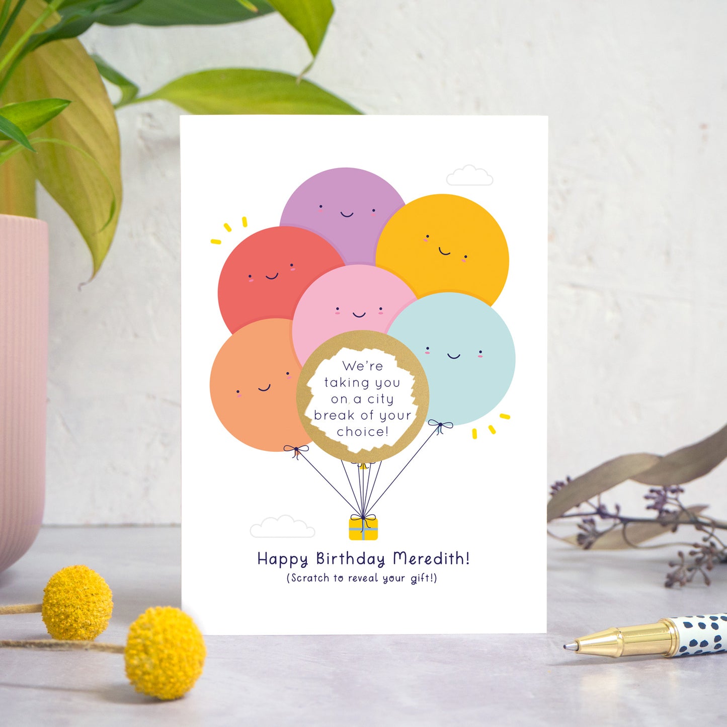 A ‘happy birthday’ scratch card photographed on a white and grey background with a plant on the left and foliage and a pen on the right. This is the rainbow version of the card after it has been scratched off.