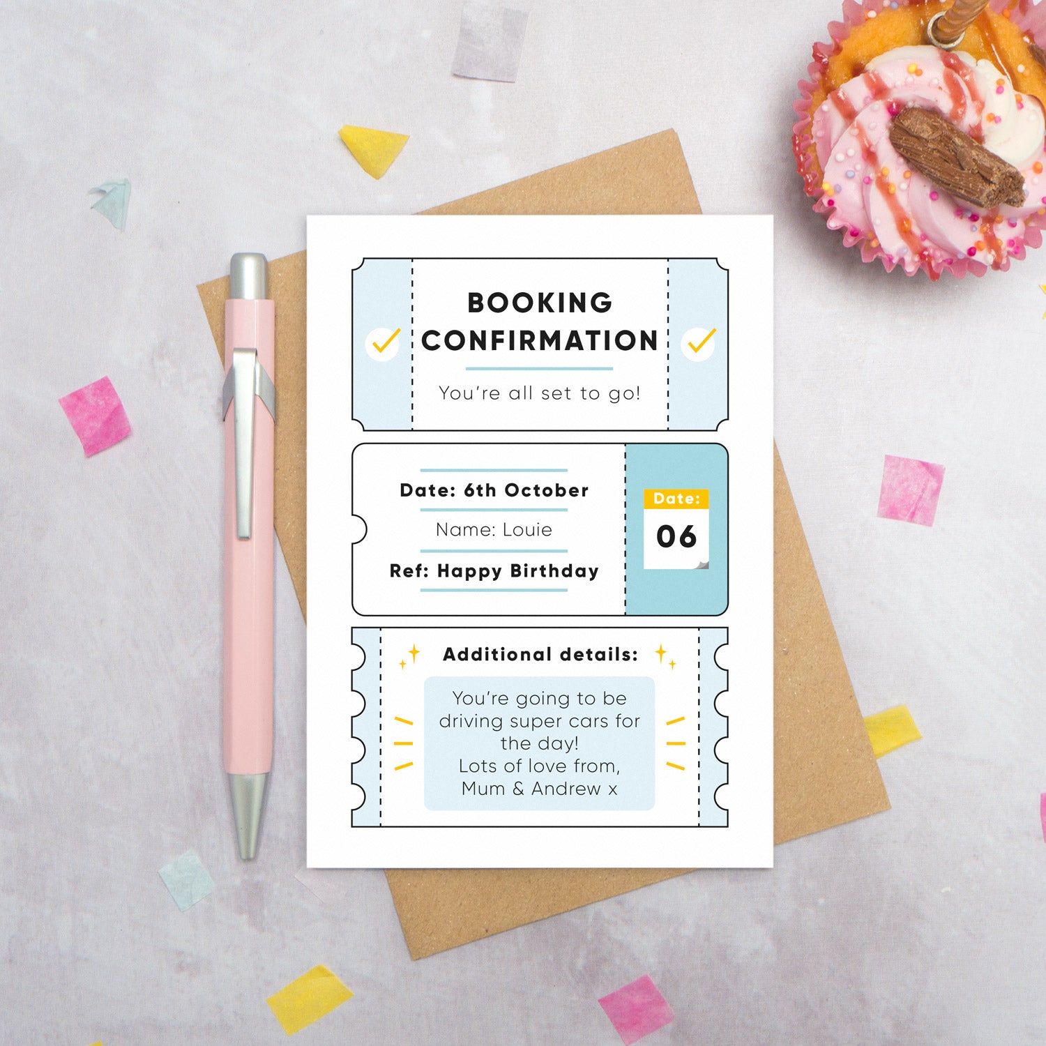 A personalised birthday booking confirmation gift card lying flat lay on a grey surface surrounded by confetti, a cupcake and a pink pen. The card is lying on it’s kraft brown envelope and pictured is the blue version of the gift card.