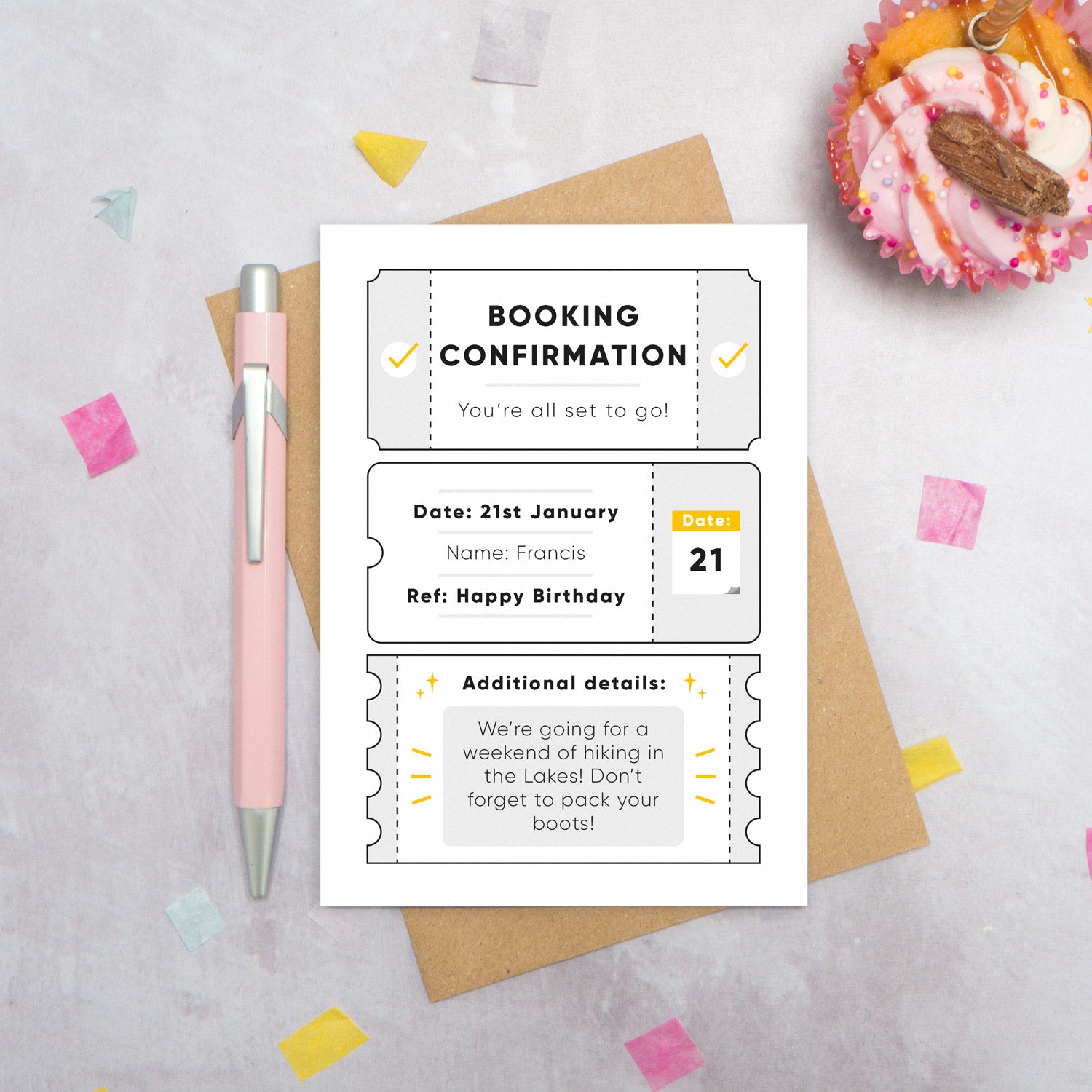 A personalised birthday booking confirmation gift card lying flat lay on a grey surface surrounded by confetti, a cupcake and a pink pen. The card is lying on it’s kraft brown envelope and pictured is the grey version of the gift card.