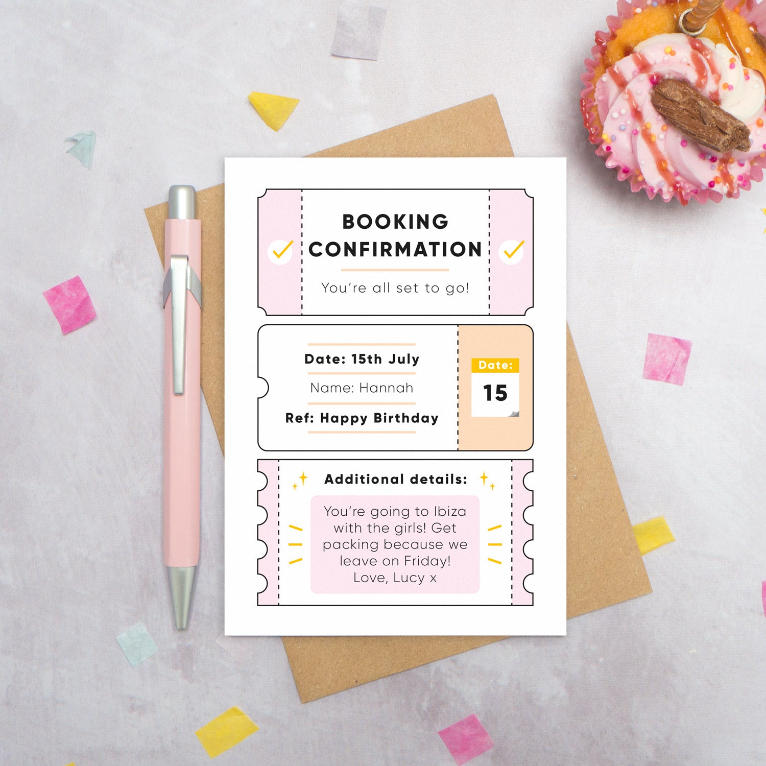 A personalised birthday booking confirmation gift card lying flat lay on a grey surface surrounded by confetti, a cupcake and a pink pen. The card is lying on it’s kraft brown envelope and pictured is the pink version of the gift card.
