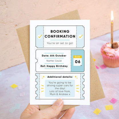 A personalised birthday booking confirmation gift card held in front of a peach and grey background scattered with birthday confetti and a cup cake with a lit candle. Pictured is the blue version of the card.