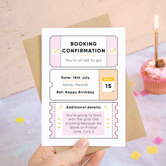 A personalised birthday booking confirmation gift card held in front of a peach and grey background scattered with birthday confetti and a cup cake with a lit candle. Pictured is the pink version of the card.