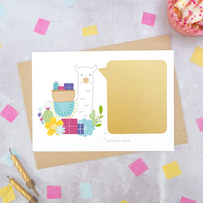 A personalised Llama birthday scratch card shot on a grey background with scattered confetti, a birthday cupcake and candles. This image is an example of how to card looks once the message has been covered with the gold scratch off panel. The card features a llama carrying gifts.