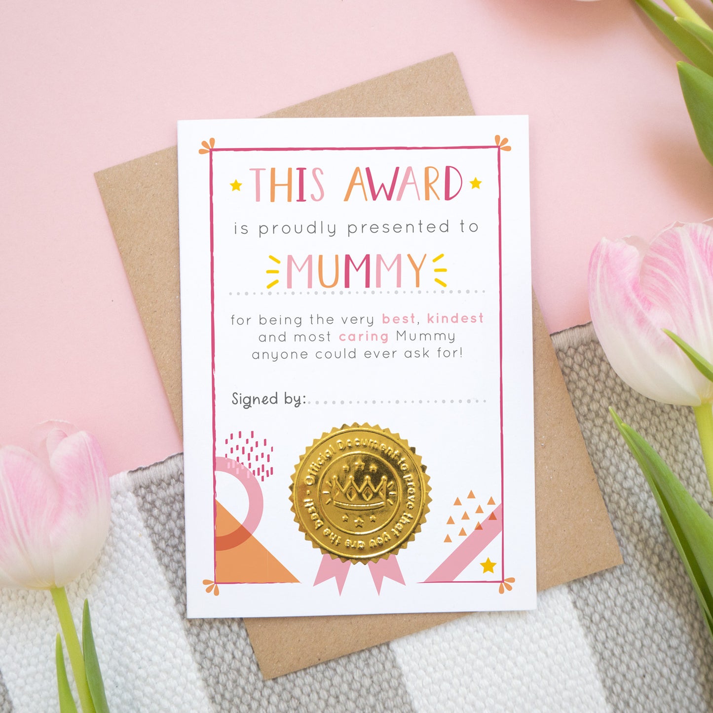 A mother's day certificate for Mummy showing the card before a child has written on the front. This is how your card will arrive. It has been shot over head on a kraft brown envelope with a pink and white and grey background with tulips.