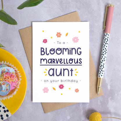 A blooming marvellous aunt birthday card photographed on top of it’s kraft brown envelope set on a grey background. To the left is a yellow plate with a birthday cupcake and to the top is green foliage. To the right is a spotty pen for scale and a pop of a yellow flower in the bottom right.