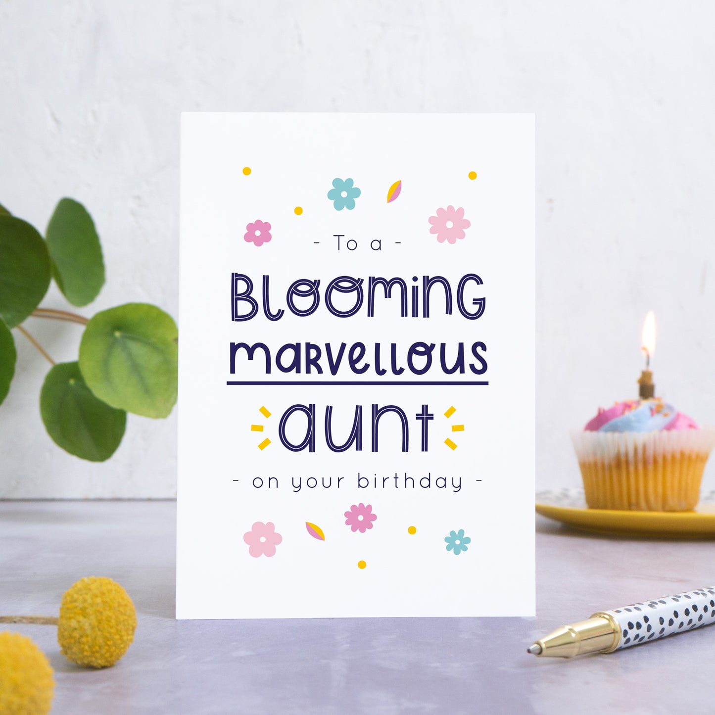 A blooming marvellous aunt birthday card photographed standing in front of a white background. In the background is a plant and a birthday cupcake and with a candle. In the foreground are two yellow flowers and a spotty pen.