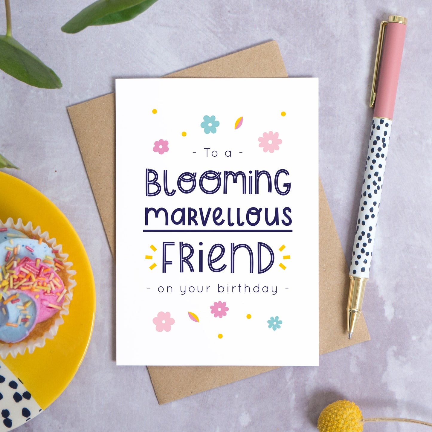 A blooming marvellous friend birthday card photographed on top of it’s kraft brown envelope set on a grey background. To the left is a yellow plate with a birthday cupcake and to the top is green foliage. To the right is a spotty pen for scale and a pop of a yellow flower in the bottom right.