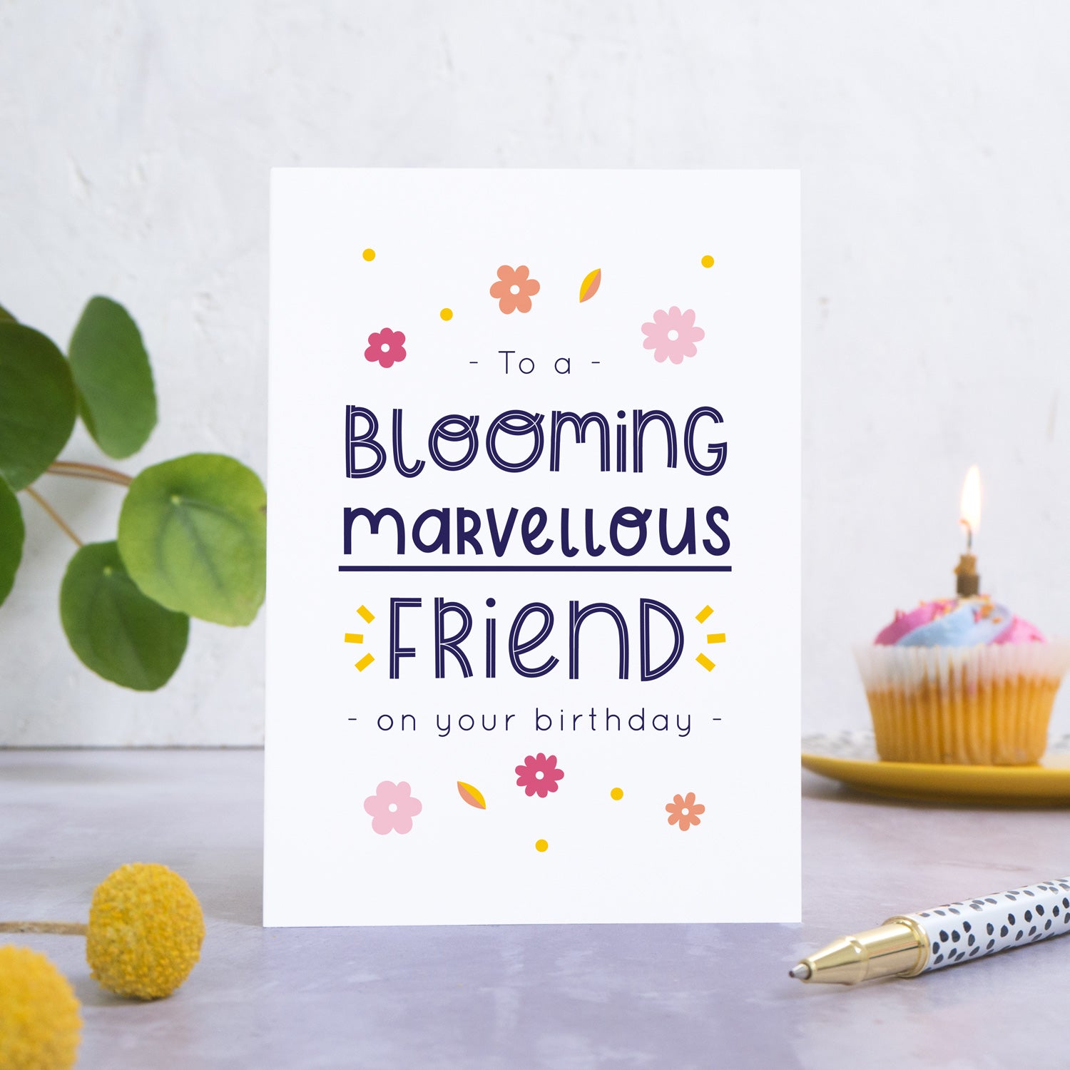 A blooming marvellous friend birthday card photographed standing in front of a white background. In the background is a plant and a birthday cupcake and with a candle. In the foreground are two yellow flowers and a spotty pen.