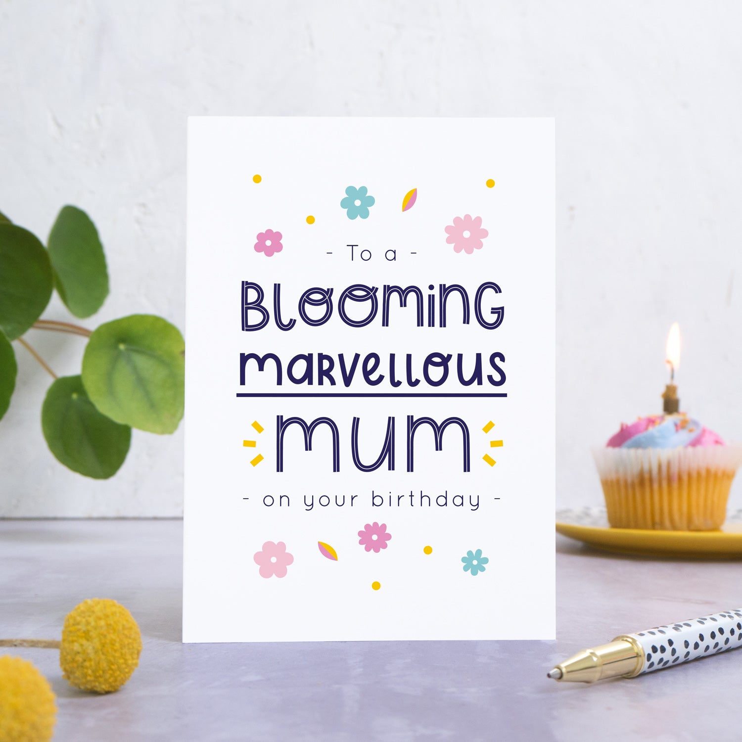 A blooming marvellous mum birthday card photographed standing in front of a white background. In the background is a plant and a birthday cupcake and with a candle. In the foreground are two yellow flowers and a spotty pen.