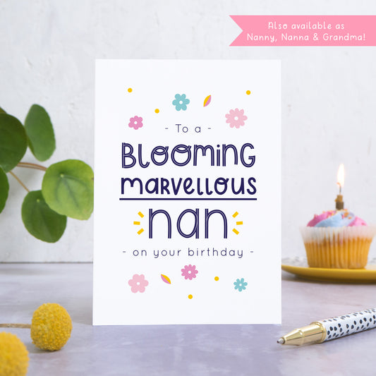 A blooming marvellous nan birthday card photographed standing in front of a white background. In the background is a plant and a birthday cupcake and with a candle. In the foreground are two yellow flowers and a spotty pen.