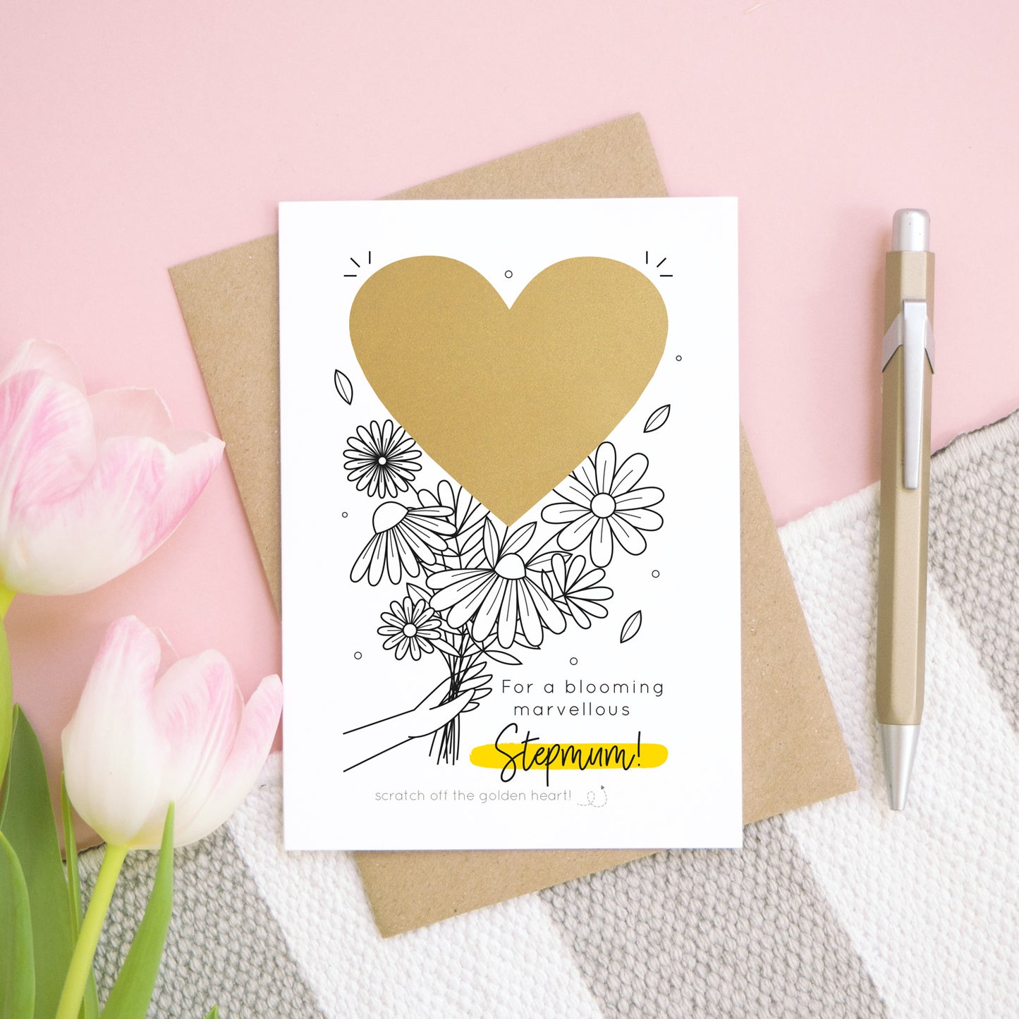 A personalised blooming marvellous scratch card lying flat on top of its kraft brown envelope on a pink, white and grey background. There are pink tulips to the left and a pen to the right for scale. The heart is still in it’s solid form hiding the scratch off message.