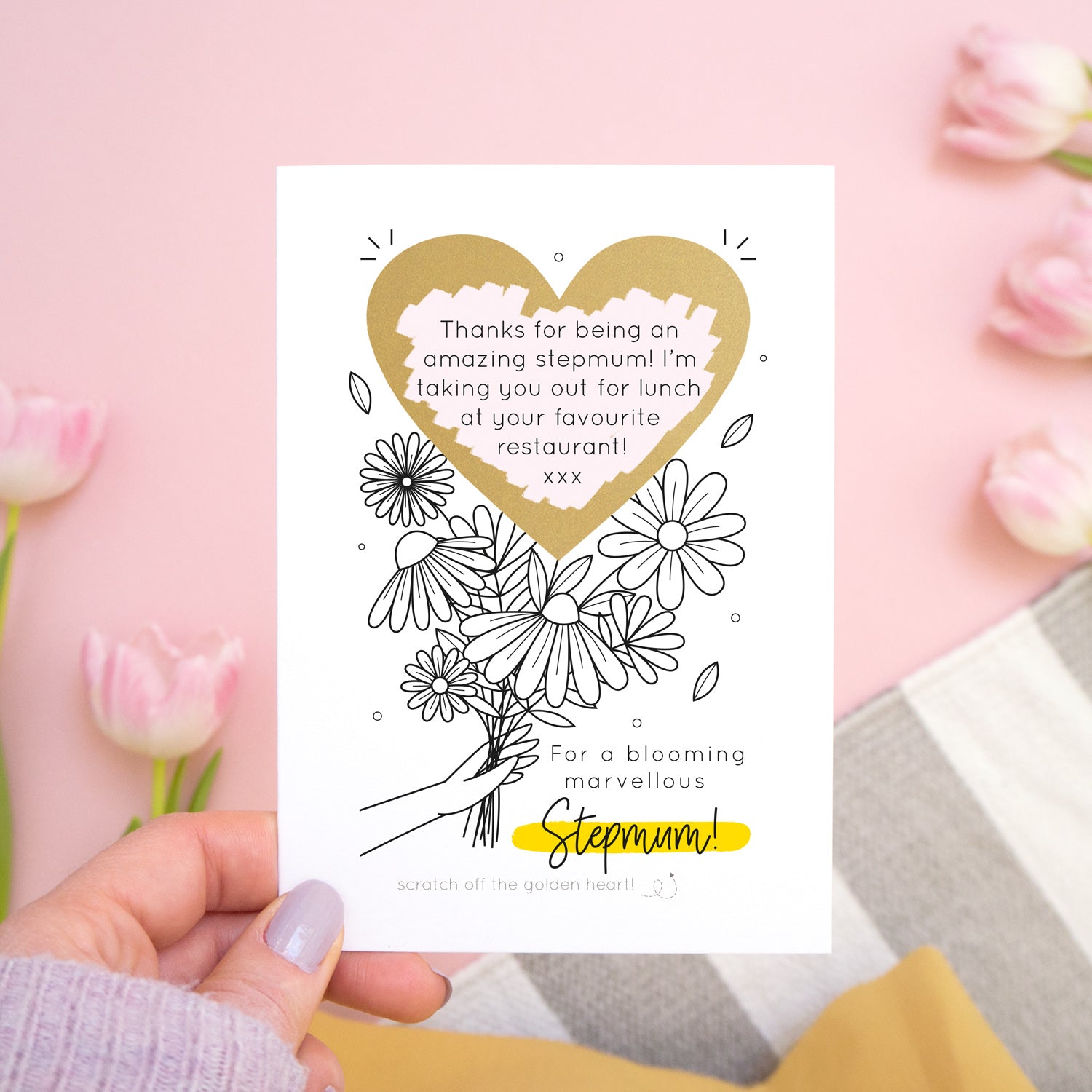 A personalised blooming marvellous scratch card being held above a pink background by someone in a yellow skirt over a grey and white rug. There are pink tulips around the edge of the photo. The card features the black and yellow colour way with the scratch off heart revealing the custom printed message.