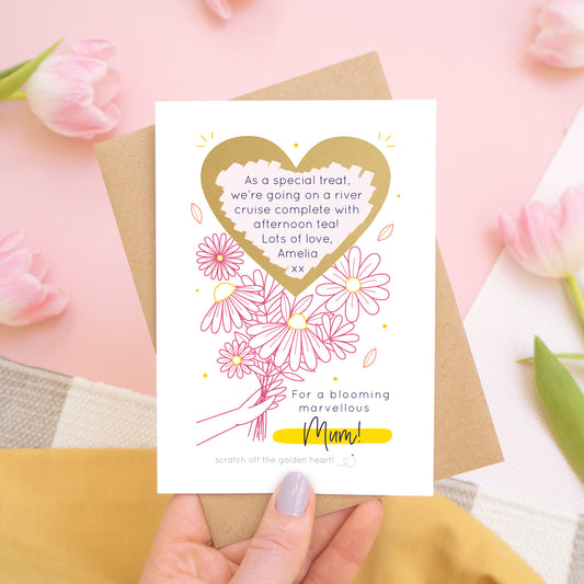 A personalised blooming marvellous scratch card being held above a pink background by someone in a yellow skirt over a grey and white rug. There are pink tulips around the edge of the photo. The card is being held against its kraft brown envelope and features the pink and yellow colour way with the scratch off heart revealing the custom printed message.