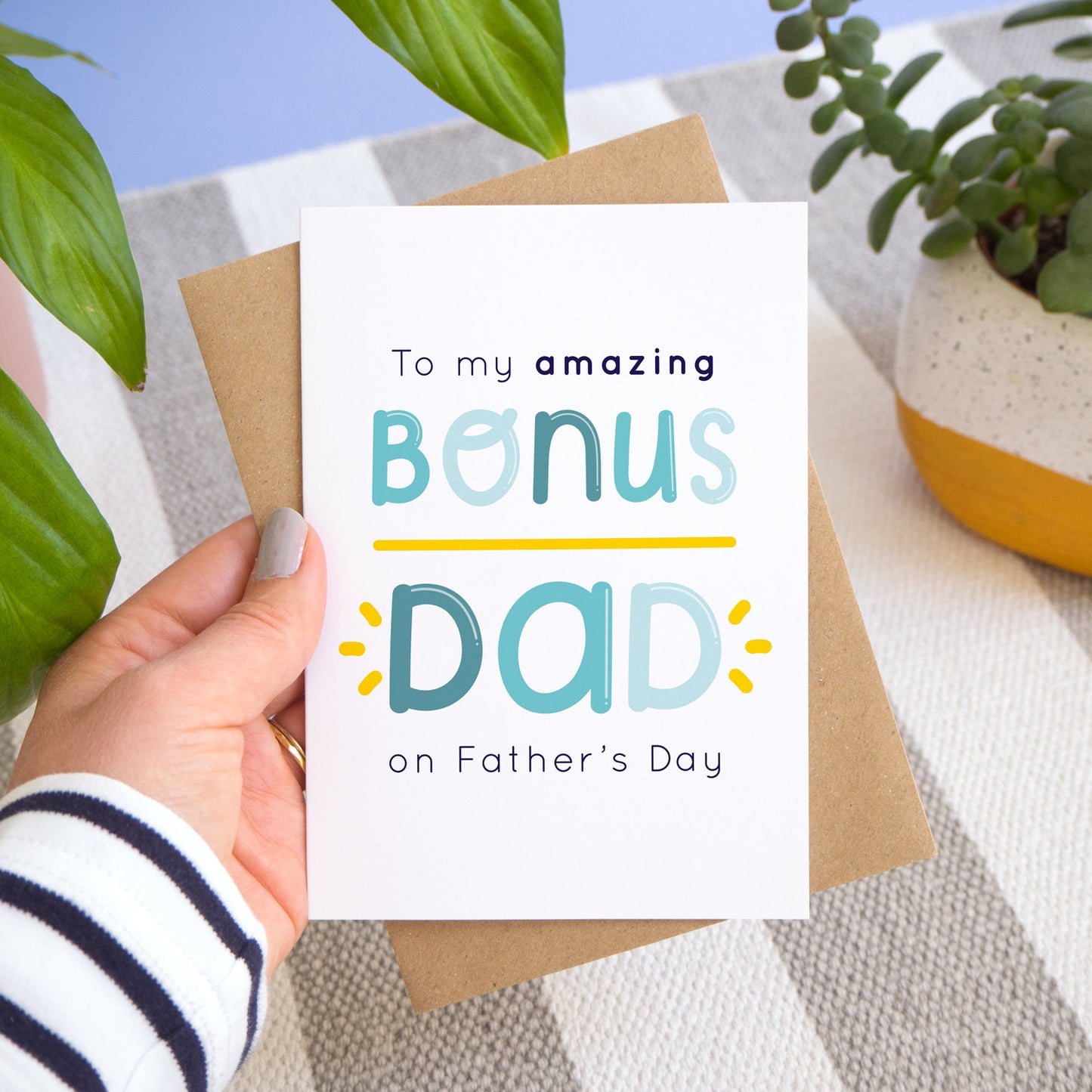The bonus dad Father's day card and kraft brown envelope are held over a stripy rug with a blue background and potted plants. The card feature navy and varying tones of blue writing and a yellow underline and flicks.