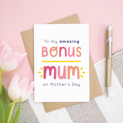 A bonus mum mother’s day card designed for your stepmum positioned on top of a kraft brown envelope on top of a over a pink background with a grey and white rug with pink tulips. This card is in the pink and peach colour palette.