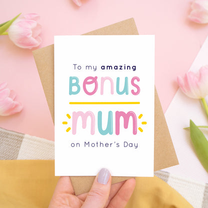 A bonus mum mother’s day card designed for your stepmum held on top of a kraft brown envelope over a pink background with pink tulips. This card is in the pink and blue colour palette.