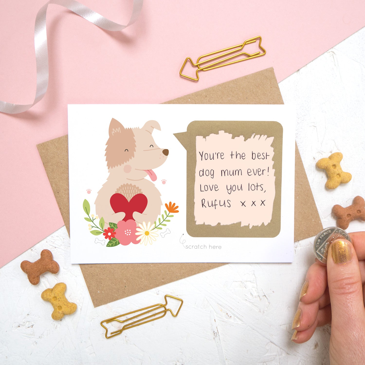 A personalised dog scratch card with a hand written message that has been scratched off. Photographed on a pink and white background with dog biscuits for props.