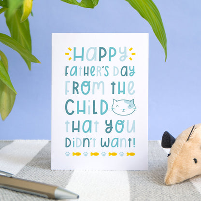 A cat child that you didn't want card photographed standing up on a stripy rug, with a blue background, pot plants and some dog treats! The card has varying tones of blue text, a cat illustration, paw prints and fish.