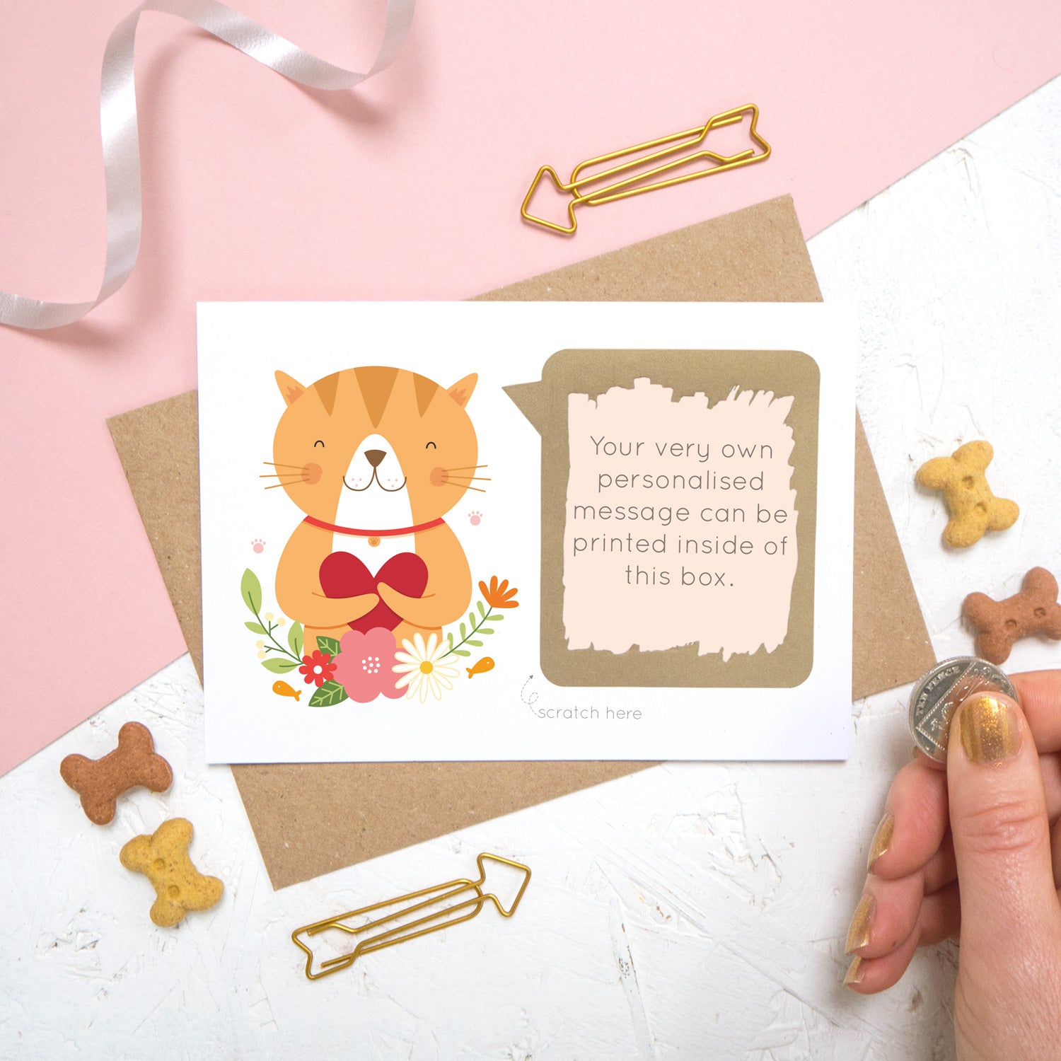 A personalised cat scratch card with the printed message revealed after it has been scratched off. The card is shot on a pink and white background with animal biscuits.