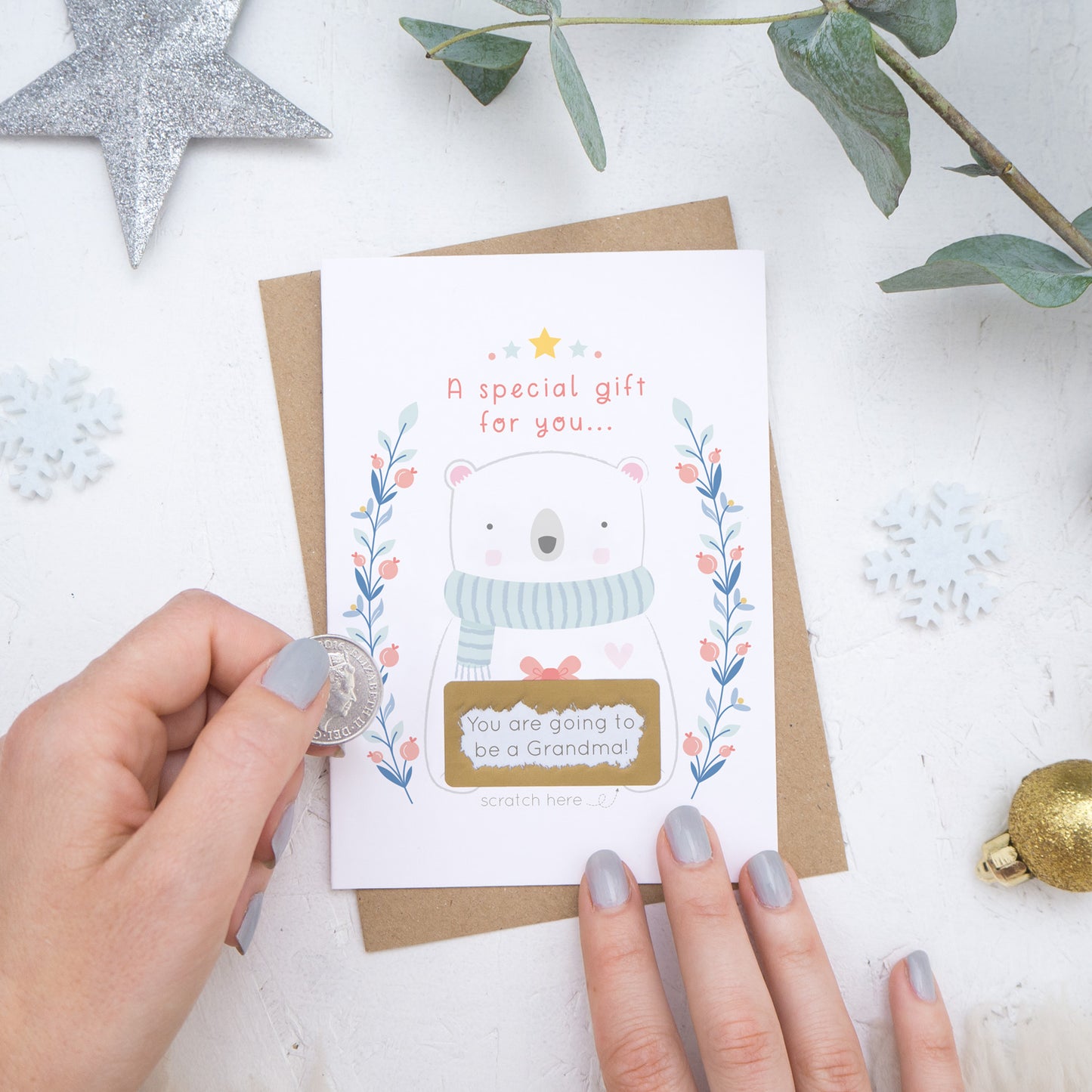 A Christmas baby announcement scratch card that has just been scratched off where the message reads 'you are going to be a grandma'. The card is on a white background surrounded with baubles, foliage and hands.