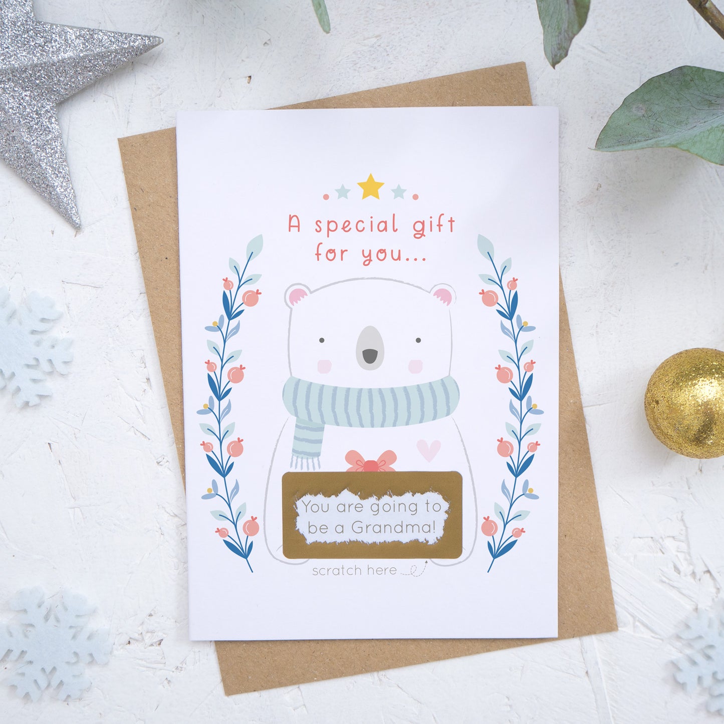 A Christmas baby announcement scratch card that has just been scratched off where the message reads 'you are going to be a grandma'. The card is on a white background surrounded with baubles, foliage and snow flakes