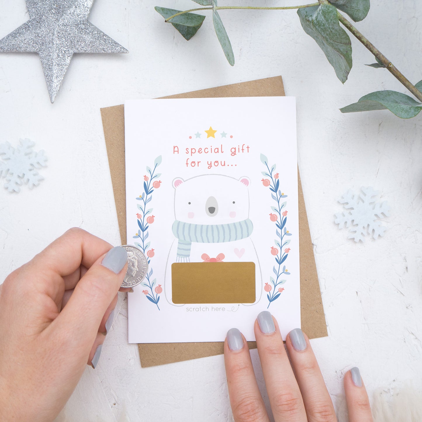 A Christmas baby announcement scratch card with a gold panel that is about to be scratched off. The card is on a white background surrounded with baubles, foliage and hands.