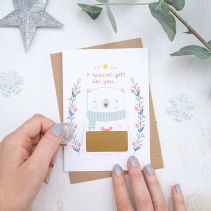 A Christmas baby announcement scratch card with a gold panel that is about to be scratched off. The card is on a white background surrounded with baubles, foliage and hands.