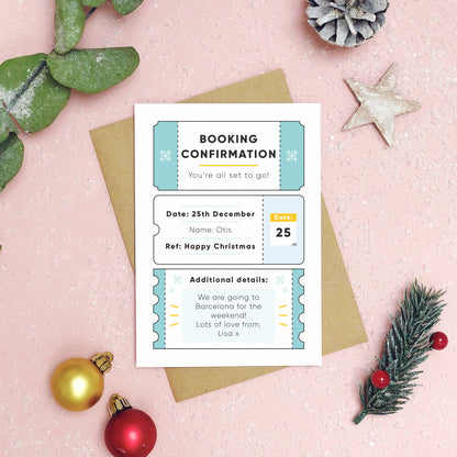 A personalised christmas booking confirmation gift card lying flat lay on a pink background scattered with foliage, baubles, and a pine cone. The card is lying on it’s kraft brown envelope and pictured is the blue version of the gift card.