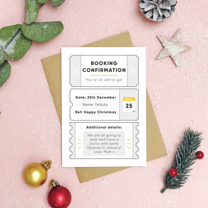 A personalised christmas booking confirmation gift card lying flat lay on a pink background scattered with foliage, baubles, and a pine cone. The card is lying on it’s kraft brown envelope and pictured is the grey version of the gift card.