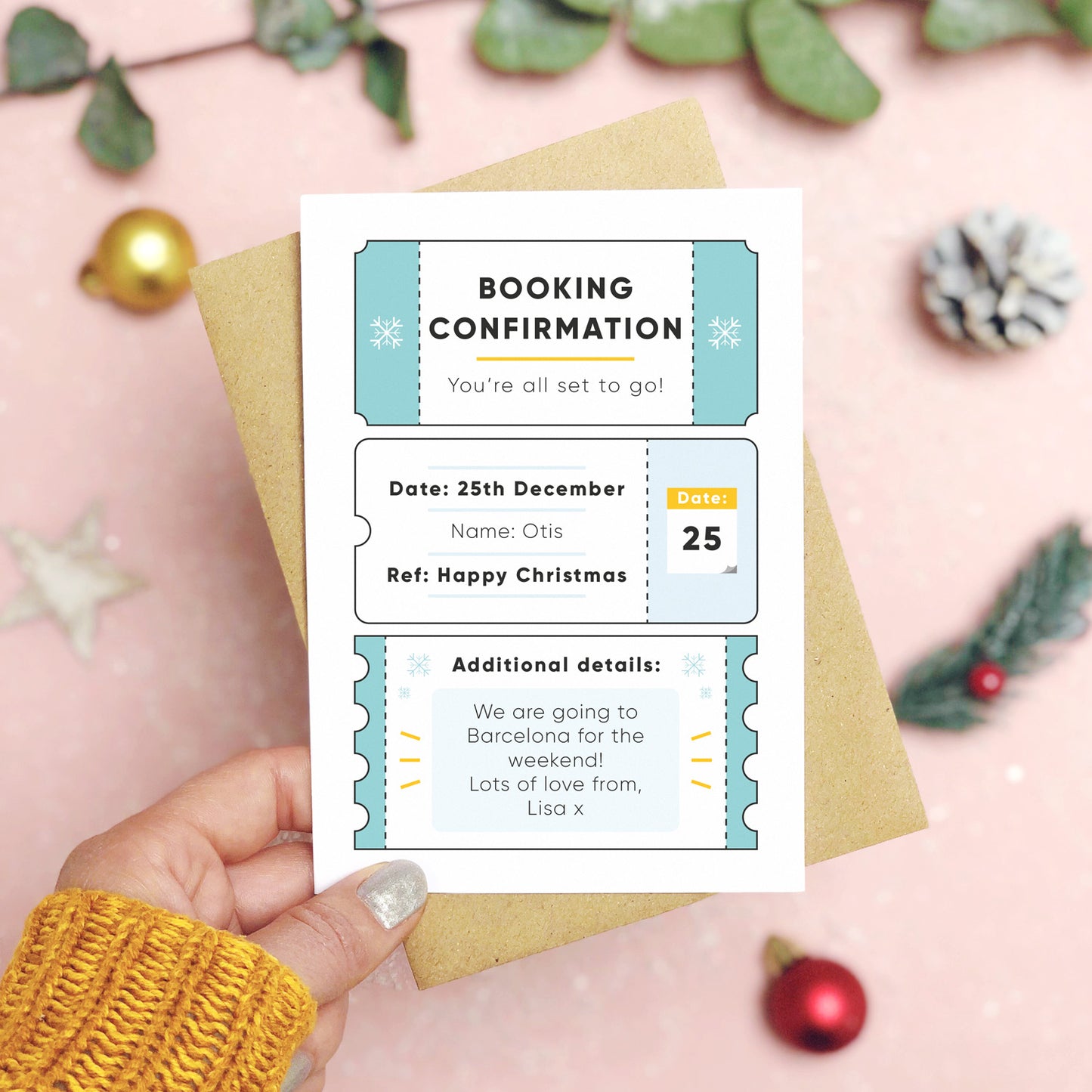 A personalised christmas booking confirmation gift card held in front of a pink background scattered with foliage, baubles, and a pine cone. Pictured is the blue version of the card on top of a kraft brown envelope.