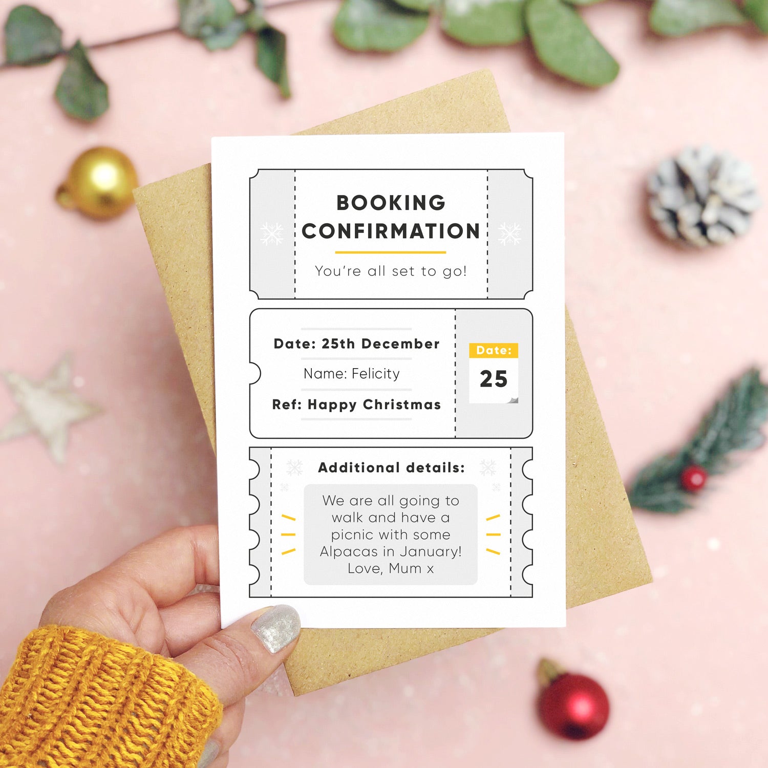A personalised christmas booking confirmation gift card held in front of a pink background scattered with foliage, baubles, and a pine cone. Pictured is the grey version of the card on top of a kraft brown envelope.