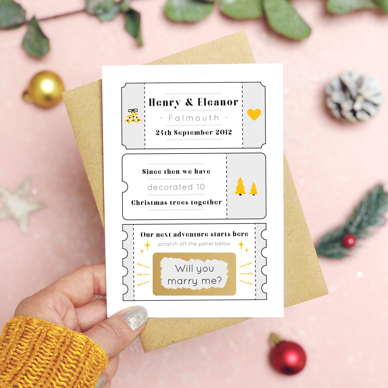A personalised will you marry me christmas scratch card in grey held over a pink background with green foliage and gold and red baubles. The card is split up into tickets and the scratch panel has been scratched off to reveal the words ‘will you marry me?’.