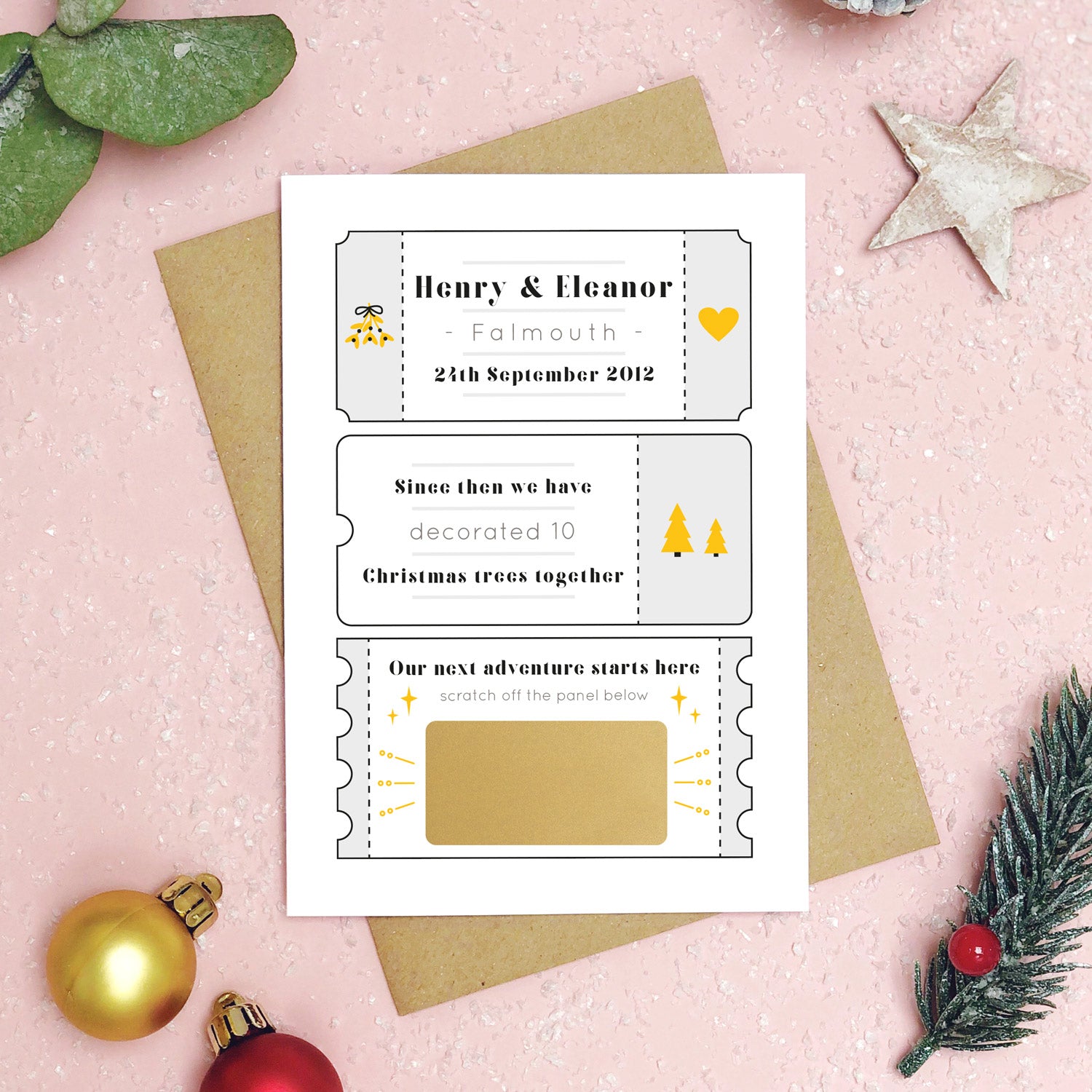 A personalised will you marry me christmas scratch card in grey lying flat a pink background with green foliage and gold and red baubles. The card is split up into tickets and the scratch panel has not yet been scratched off!