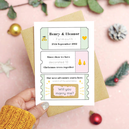 A personalised will you marry me christmas scratch card in pink and green held over a pink background with green foliage and gold and red baubles. The card is split up into tickets and the scratch panel has been scratched off to reveal the words ‘will you marry me?’.