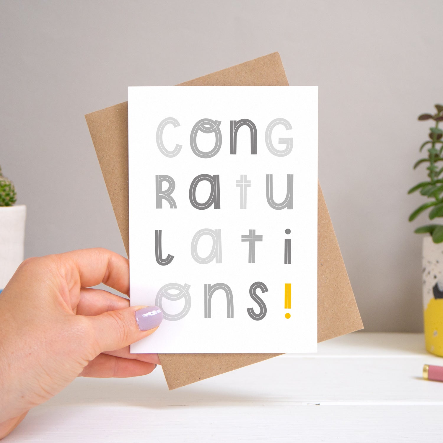 A congratulations card held over a warm grey and white background with potted plants peeping the sides. Behind the card is a kraft brown envelope that comes with the card. The text on this version of the card is in varying tones of grey.