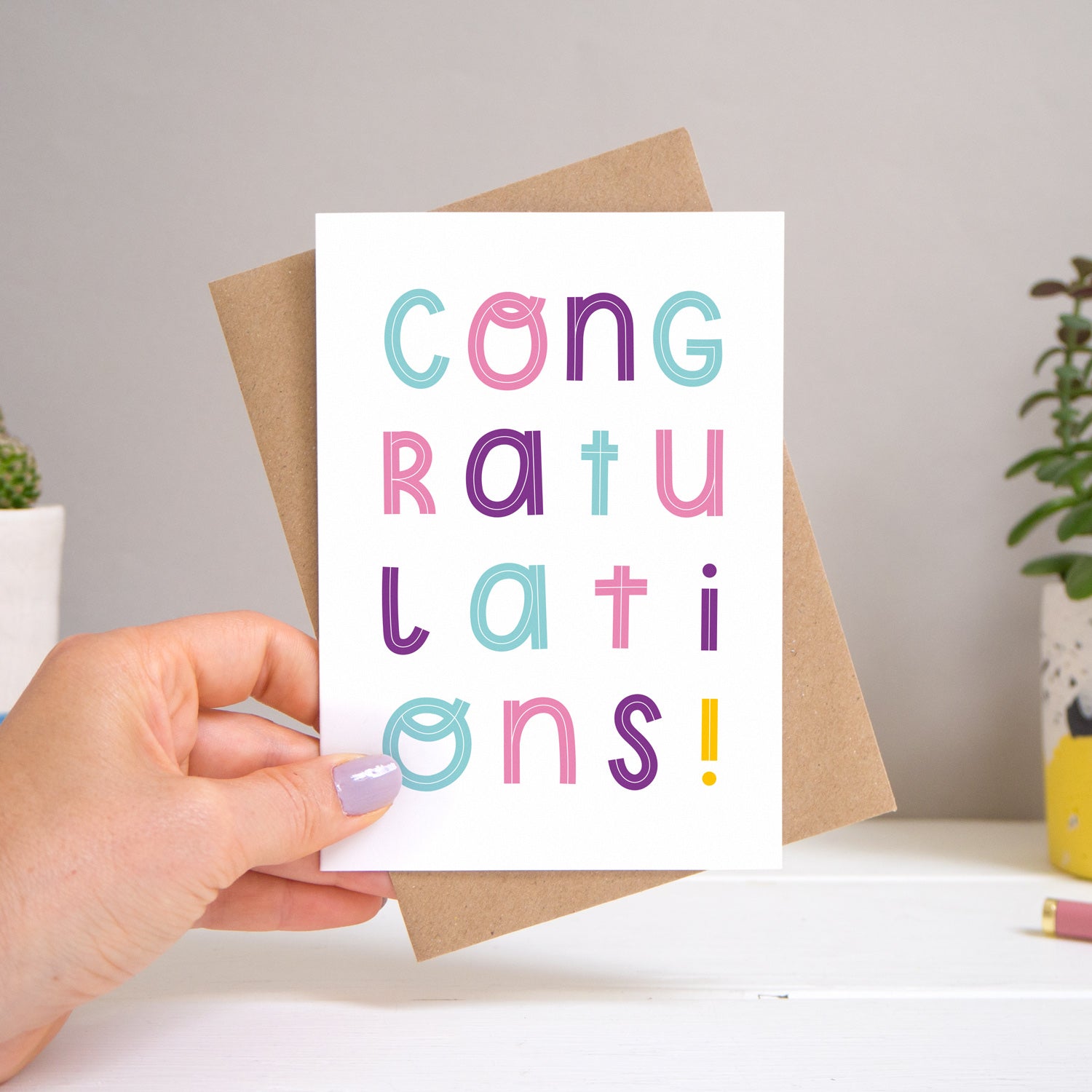 A congratulations card held over a warm grey and white background with potted plants peeping the sides. Behind the card is a kraft brown envelope that comes with the card. The text on this version of the card is in varying tones of pink, purple and blue.
