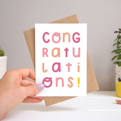 A congratulations card held over a warm grey and white background with potted plants peeping the sides. Behind the card is a kraft brown envelope that comes with the card. The text on this version of the card is in varying tones of pink.