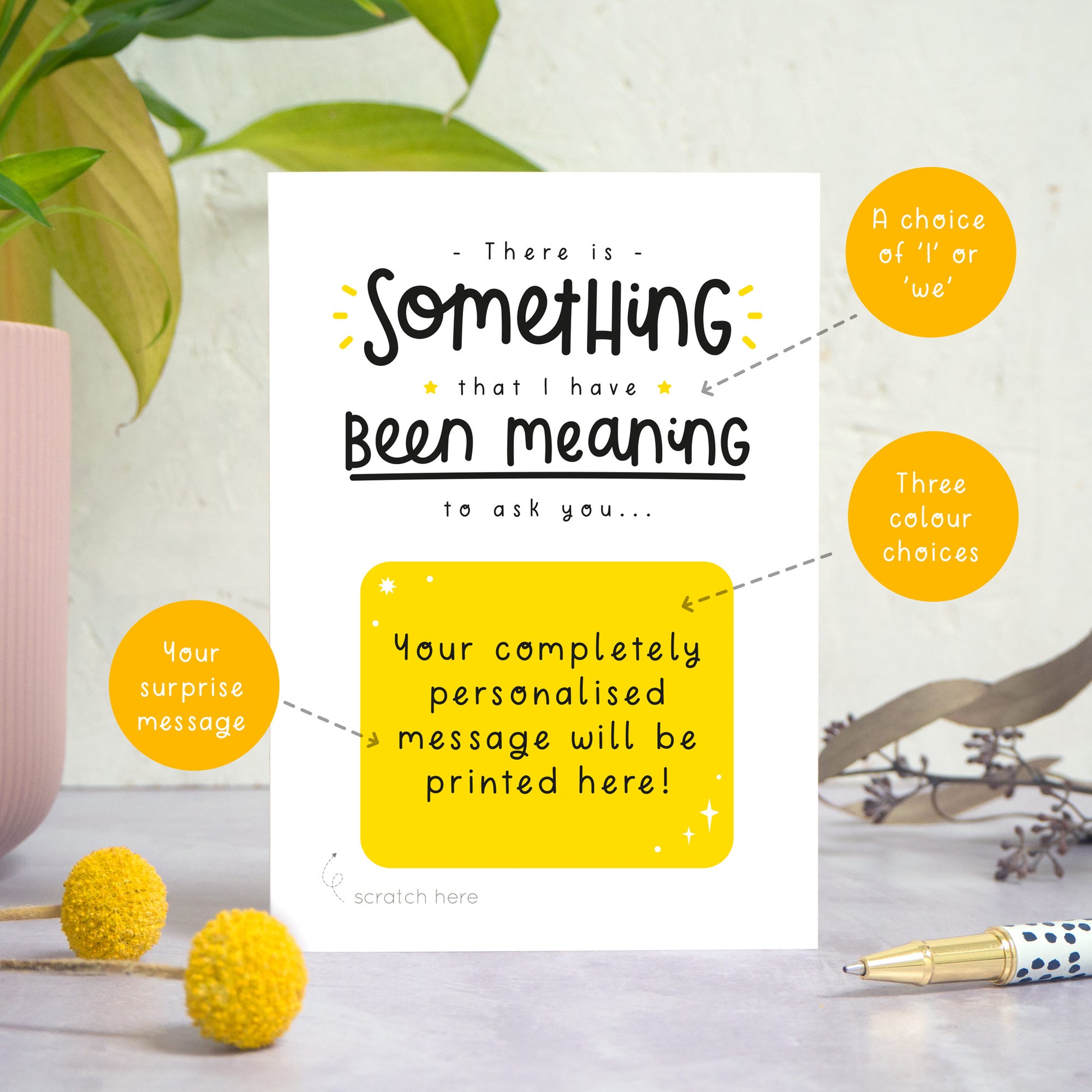 A personalised custom question scratch card photographed stood on a grey surface, a white textured background and with a pot plant on the left. Some small flowers and a pen are also in the foreground. This card is the black and yellow colour way. The orange circles show areas of the card that contain choices e.g. colour, wording and scratch message.