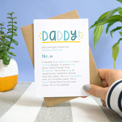A daddy definition card being held over a stripy carpet and a blue background with potted plants behind. The card shows daddy in bold lettering with the personalised nicknames underneath, followed by the definition.