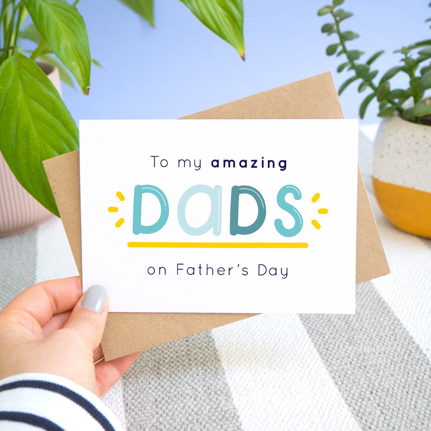 An amazing dads card being held over a stripy carpet and a blue background with potted plants behind. The card shows navy and varying tones of blue text with a yellow underline and flicks. The card reads: "to my amazing dads on Father's day"