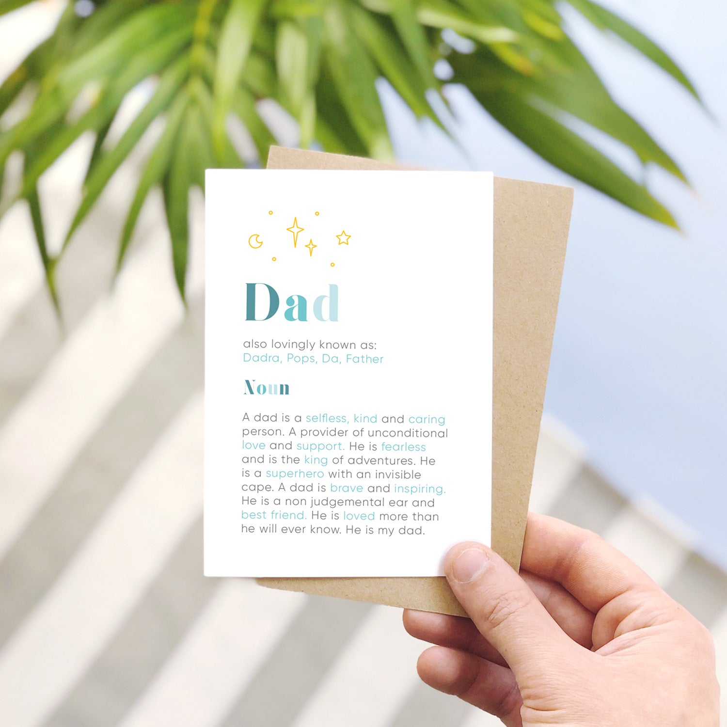 A personalised Dad dictionary definition card, shot on a grey stripy and blue background. The card features a definition of a dad in grey and blue text with yellow stars and dots.